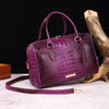 A deep purple crocodile pattern luxury leather crossbody tote bag resting on a light surface next to a piece of driftwood.