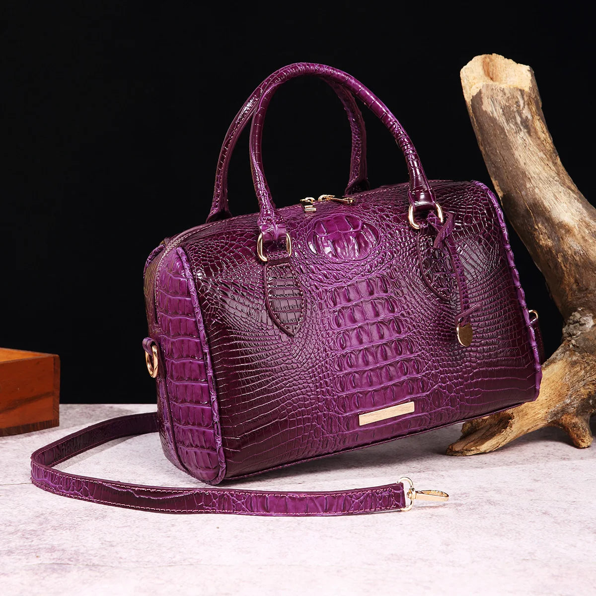 A deep purple crocodile pattern luxury leather crossbody tote bag resting on a light surface next to a piece of driftwood.