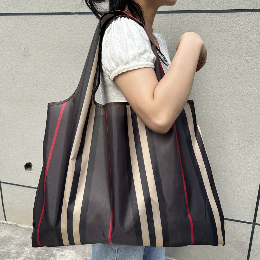 A person is holding a large, striped black and beige grocery tote bag against a light gray wall.