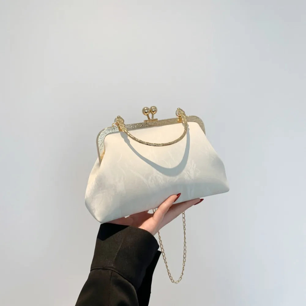 Una mano sostiene un bolso de mano blanco con lazo brillante dorado contra un fondo claro.
