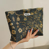 A hand holds a dark floral canvas makeup bag with gold accents against a light background.