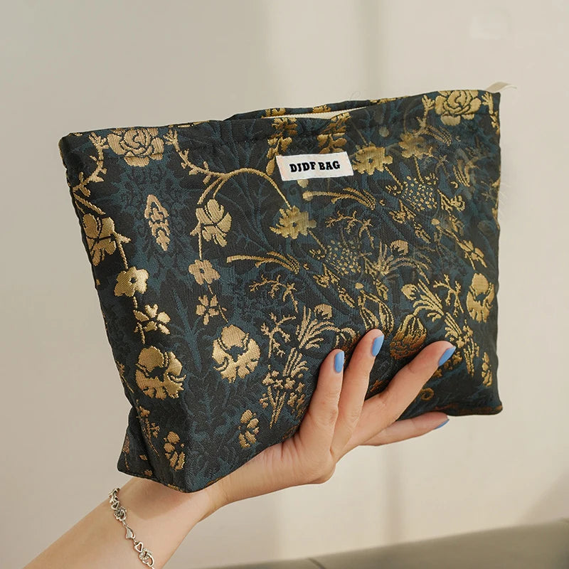 A hand holds a dark floral canvas makeup bag with gold accents against a light background.