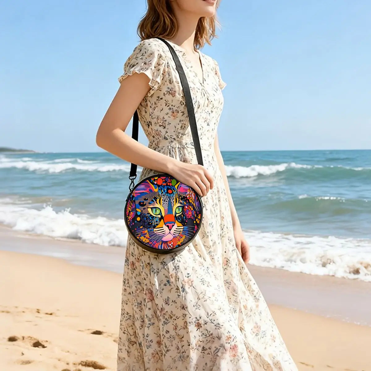 A woman wearing a floral dress holds a round, colorful cartoon cat crossbody bag on a beach.