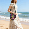 A woman wearing a floral dress holds a round, colorful cartoon cat crossbody bag on a beach.