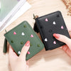 A hand holds a dark green compact PU leather tassel wallet with a zipper closure and small heart embellishments on a wooden s