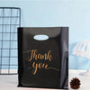 A black plastic gift bag with gold script text that reads Thank you is displayed next to a wire grid and a pine cone.