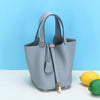 Helles graues, echtes Leder-Bucket-Bag mit Klappverschluss und goldfarbenen Details vor blauem Hintergrund.