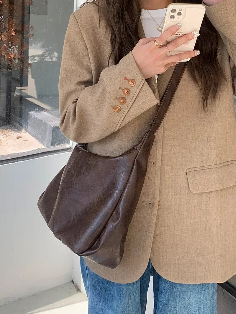 A woman holds a brown vintage PU leather tote bag with a dark brown crossbody strap against a light background.