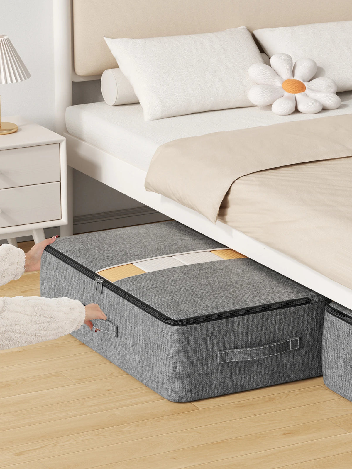 A gray foldable underbed storage bag is placed on a wooden floor next to a white bed frame.