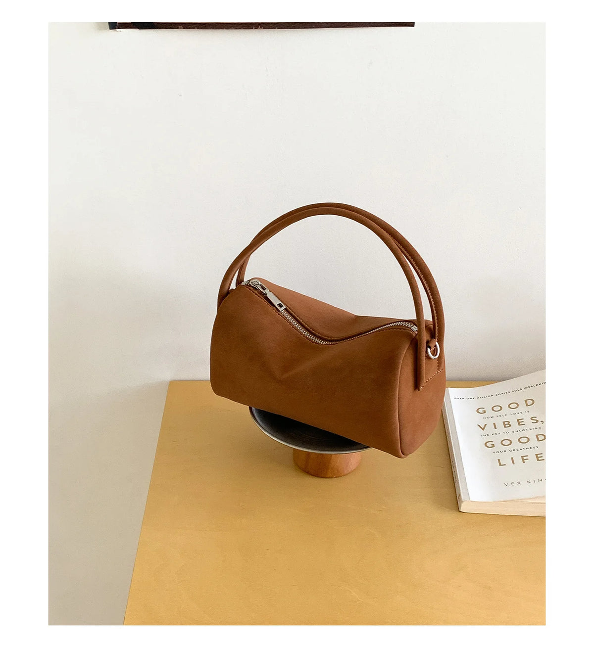 A tan faux suede crossbody bag resting on a light wood surface next to a book.