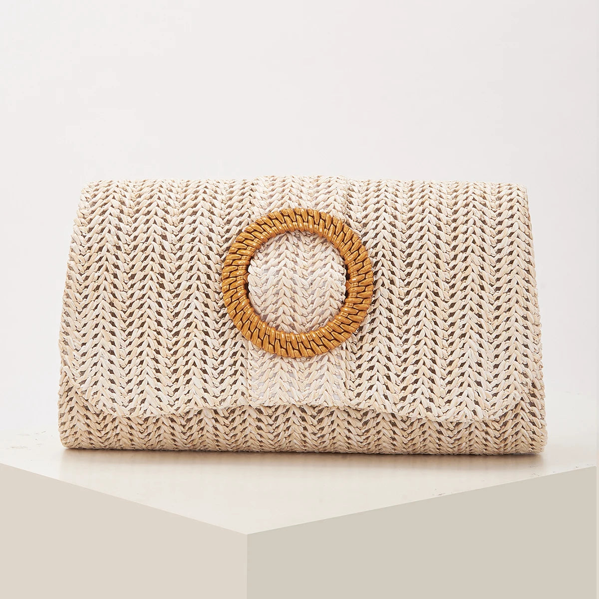 A beige woven straw envelope clutch with a round leather detail sits on a white surface.