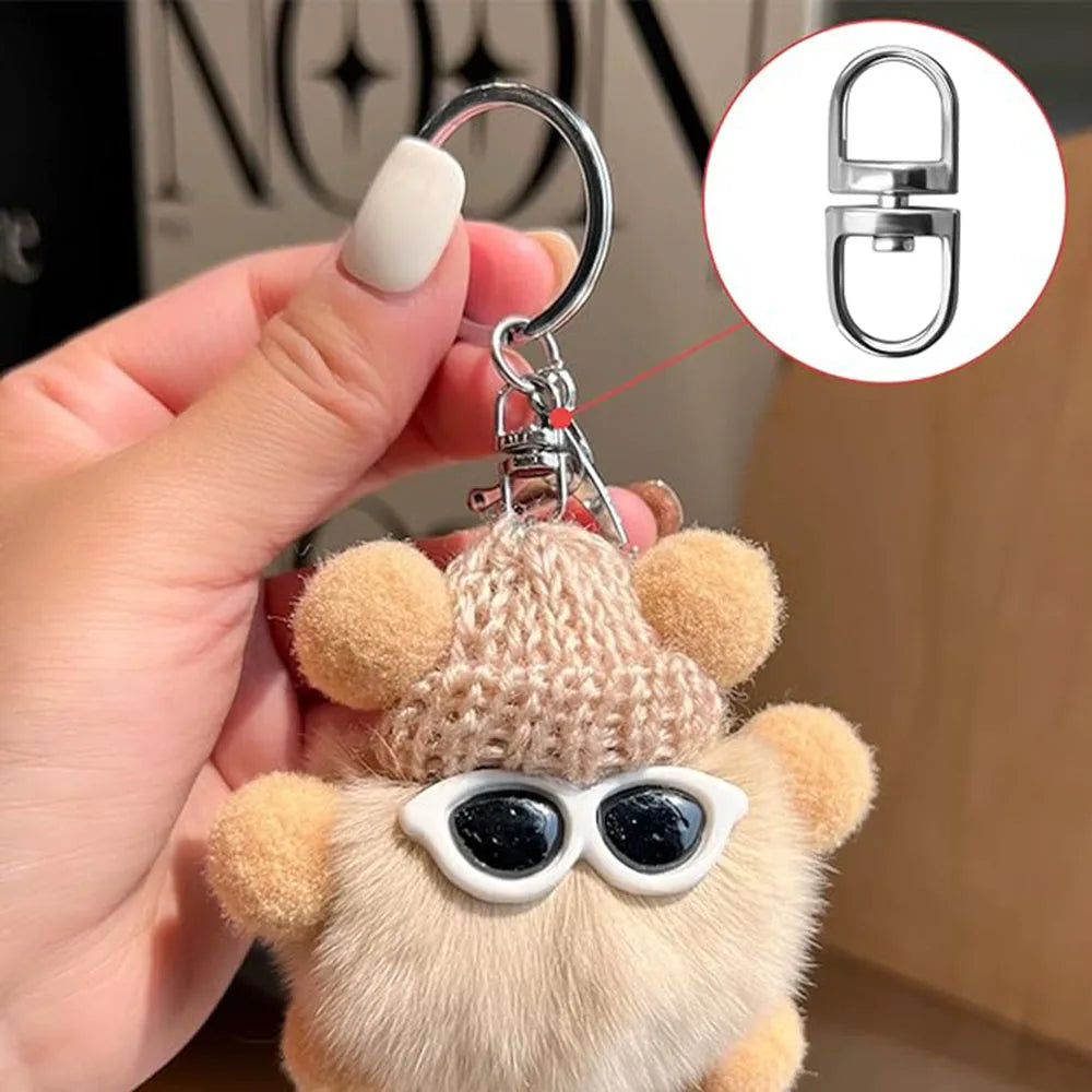 A hand holds a plush bear keychain featuring round sunglasses and swivel metal hooks.