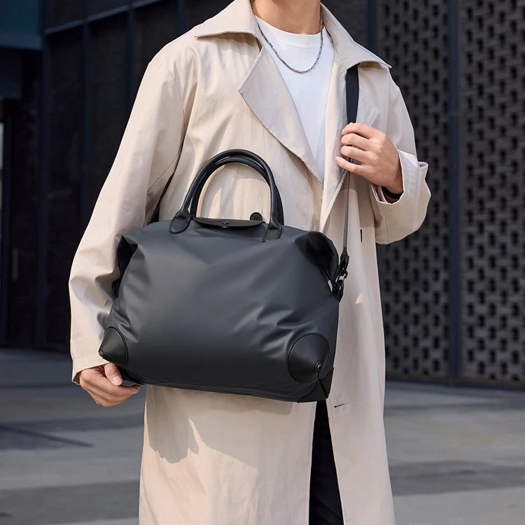 A person wearing a beige trench coat is holding a black durable nylon travel duffle bag.