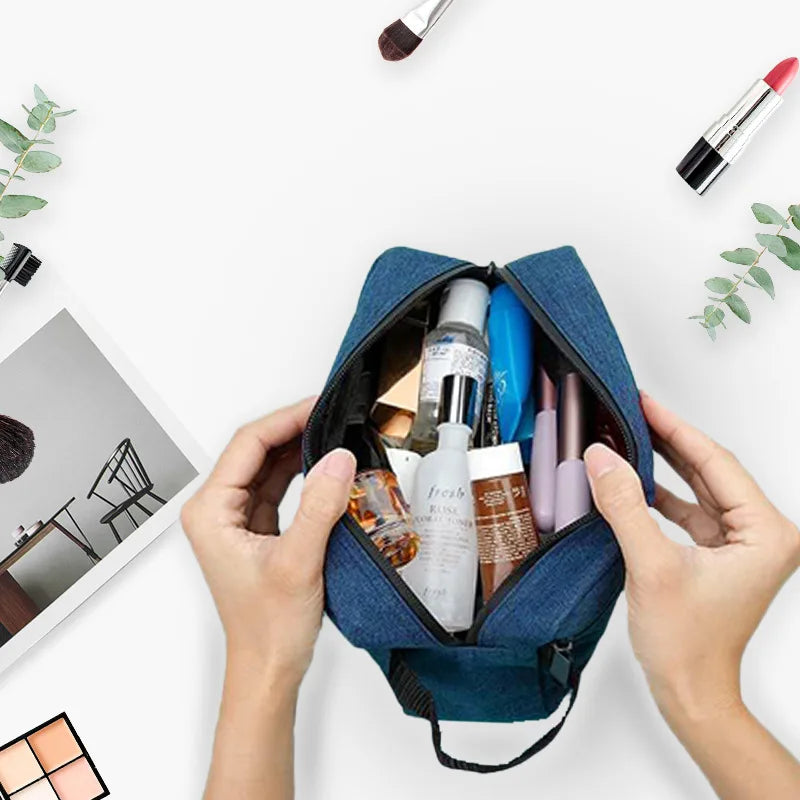 A hand holds a dark blue embroidered travel cosmetic bag filled with various makeup and beauty products.