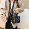 A woman models a black diamond grid textured crossbody bag with gold hardware and a beige blazer and black skirt.