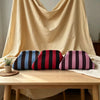 A set of three striped knit makeup bags in navy blue, maroon, and pink resting on a light wooden table.