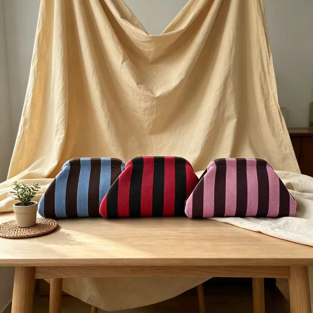 A set of three striped knit makeup bags in navy blue, maroon, and pink resting on a light wooden table.