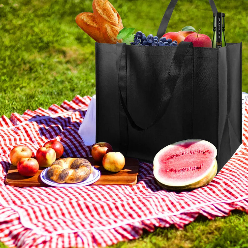 A black reusable tote bag filled with fresh fruit and bread rests on a red and white checkered blanket outdoors.