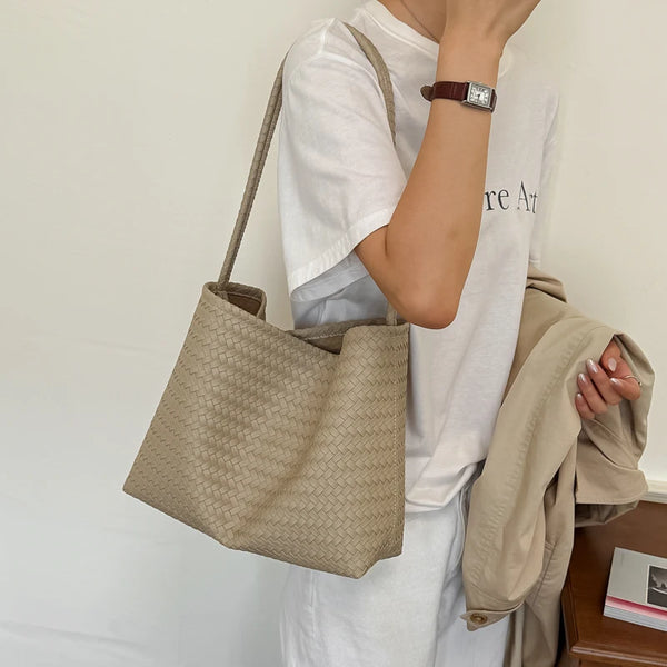 A person holds a beige PU bucket shoulder bag against a white shirt and light pants.
