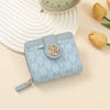 A light blue compact wallet with a gold logo and vintage letter print is shown next to a clock and a yellow tulip.