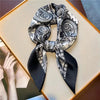 A black and white paisley silk hair scarf tied in a bow, displayed on a light wooden surface.