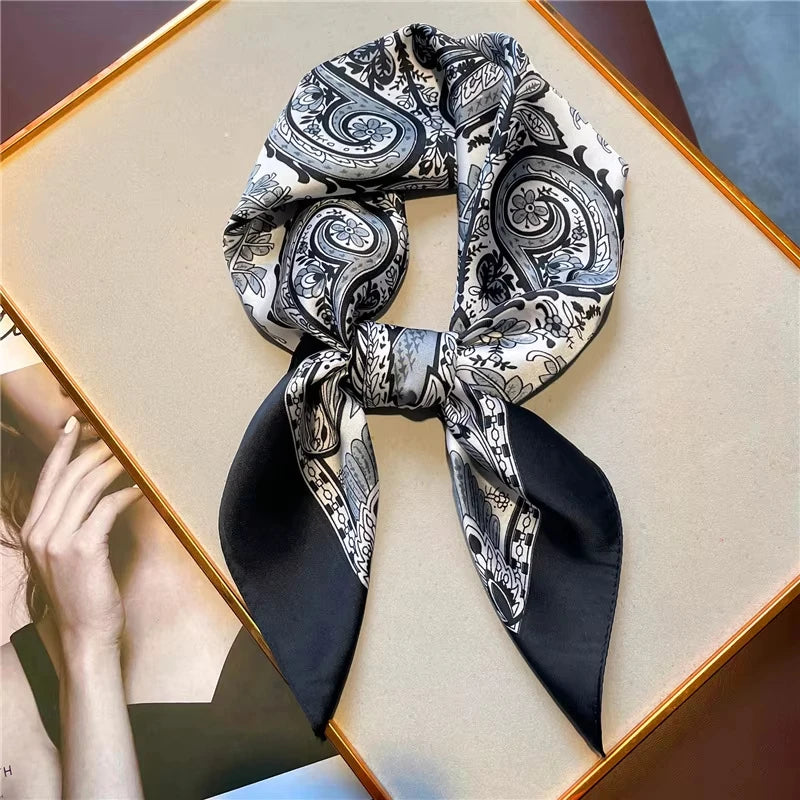 A black and white paisley silk hair scarf tied in a bow, displayed on a light wooden surface.