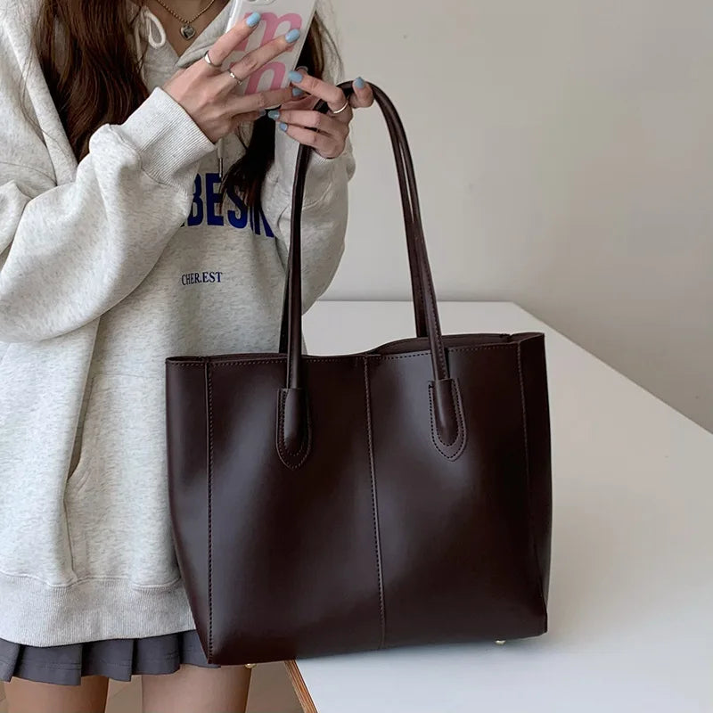 A woman holds a dark brown leather tote bag with handles against a white background.