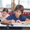 A young boy is writing at a desk with a fish-shaped pencil case and coin purse visible on the table.