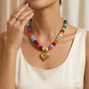 A woman wears a colorful bead necklace with resin accents and a gold heart charm against a light background.