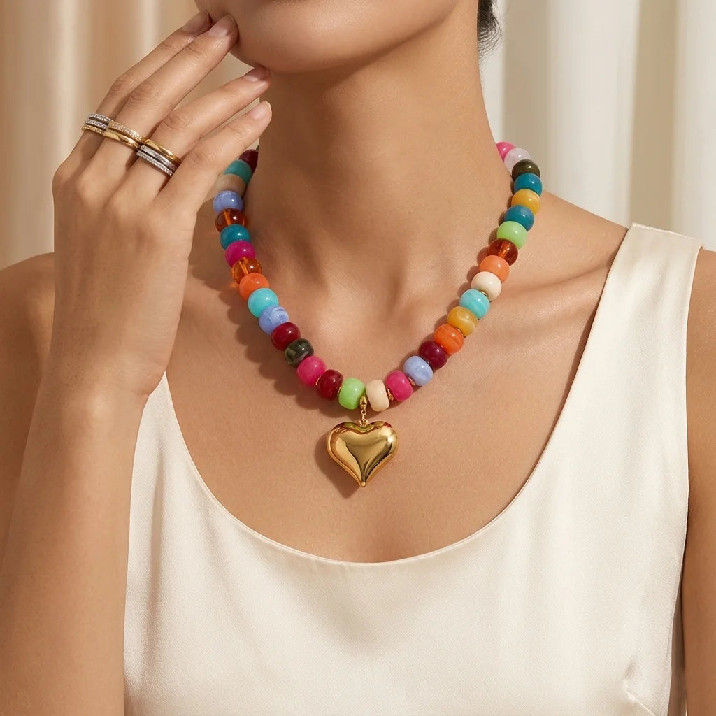 A woman wears a colorful bead necklace with resin accents and a gold heart charm against a light background.