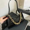 A black crocodile print shoulder bag with a gold chain strap is being held by a hand on a dark surface.