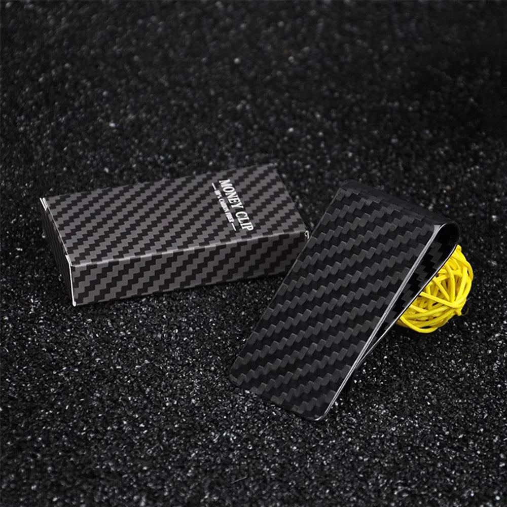 A black carbon fiber money clip is shown next to a yellow cord on a dark, textured surface.