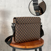 A brown patterned flap crossbody bag with a black strap displayed on a wooden stool.