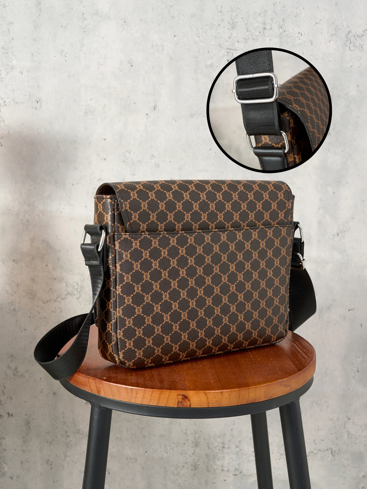 A brown patterned flap crossbody bag with a black strap displayed on a wooden stool.
