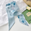 Foulard de cheveux floral en soie bleu clair noué en nœud, posé sur du papier blanc à côté d'un livre vert.