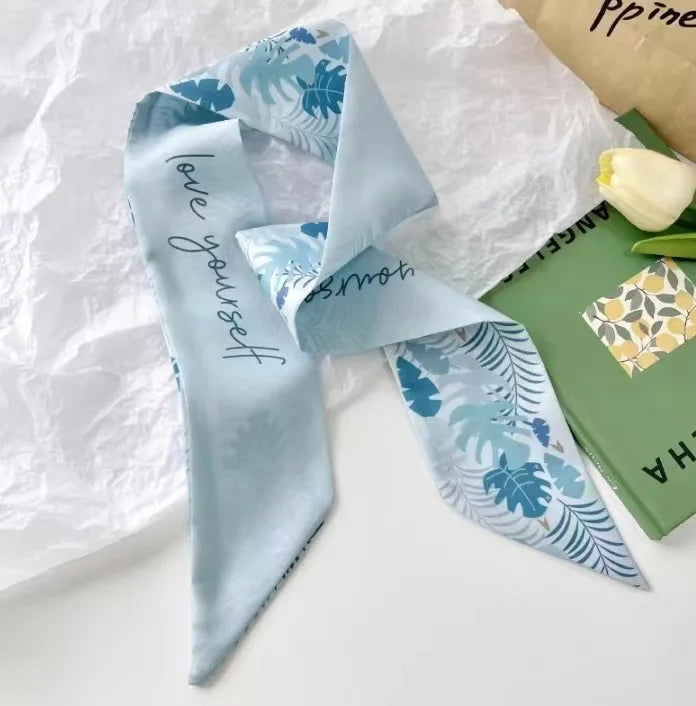 Foulard de cheveux floral en soie bleu clair noué en nœud, posé sur du papier blanc à côté d'un livre vert.