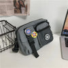 A gray canvas shoulder bag with black straps and a yellow circular tag is displayed next to a wire basket.