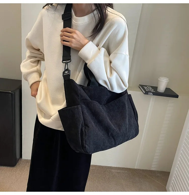 A person is holding a black corduroy multi-pocket tote bag over a cream-colored sweatshirt.