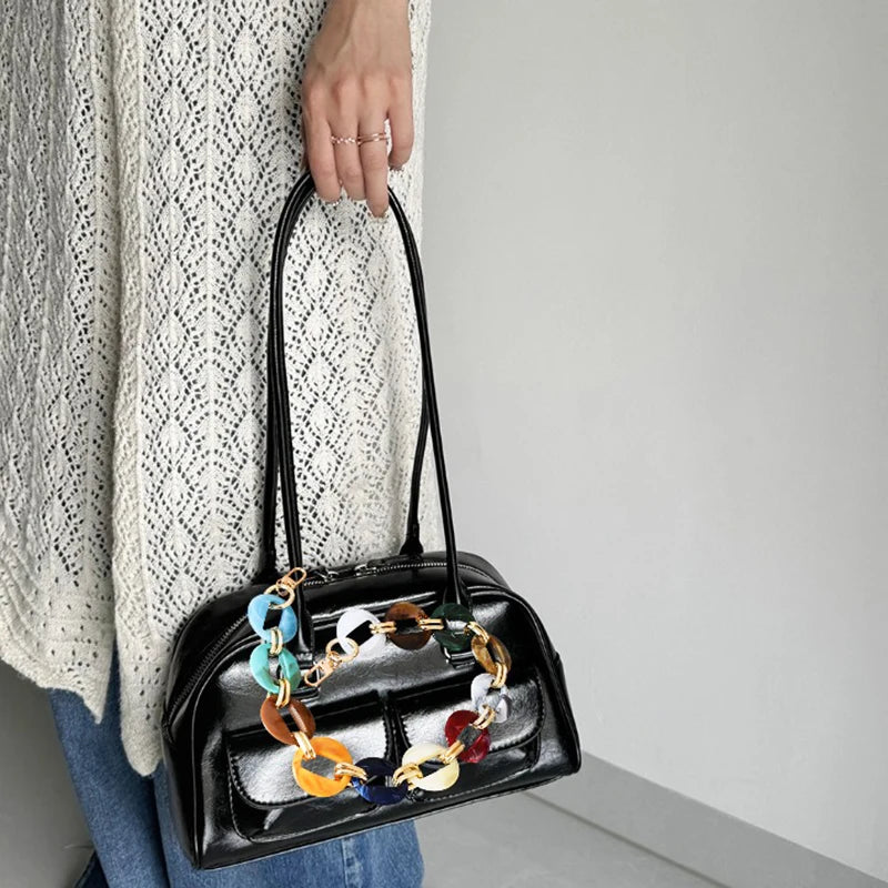 A black handbag with a multi-color acrylic chain strap is held by a person wearing a cream knit sweater.