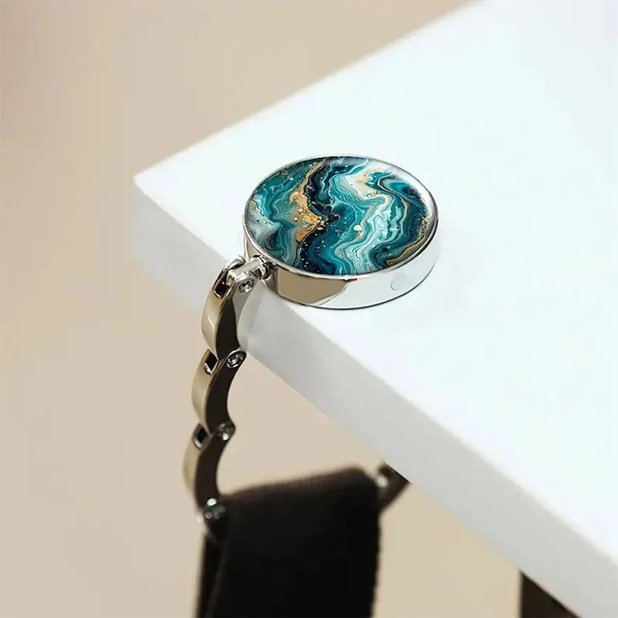 A silver-toned bag hook featuring a round pendant with a swirling blue and green marble pattern.
