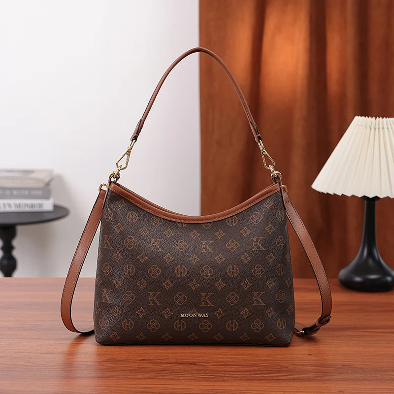 A brown leather crescent tote bag with a gold chain strap and a repeating letter print on a wooden table.