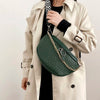 A woman wears a beige trench coat and carries a dark green woven pattern waist bag with a gold chain.