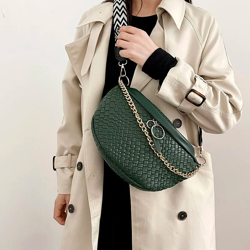 A woman wears a beige trench coat and carries a dark green woven pattern waist bag with a gold chain.