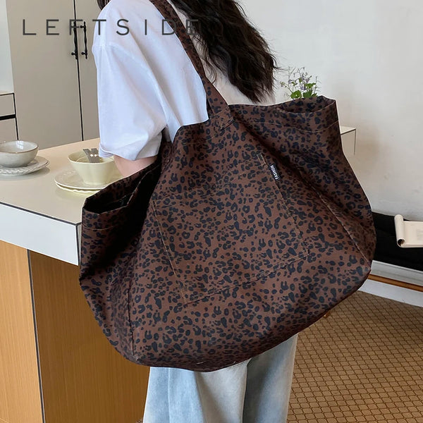 A brown leopard print canvas tote bag is shown hanging on a person's shoulder in a bright indoor setting.