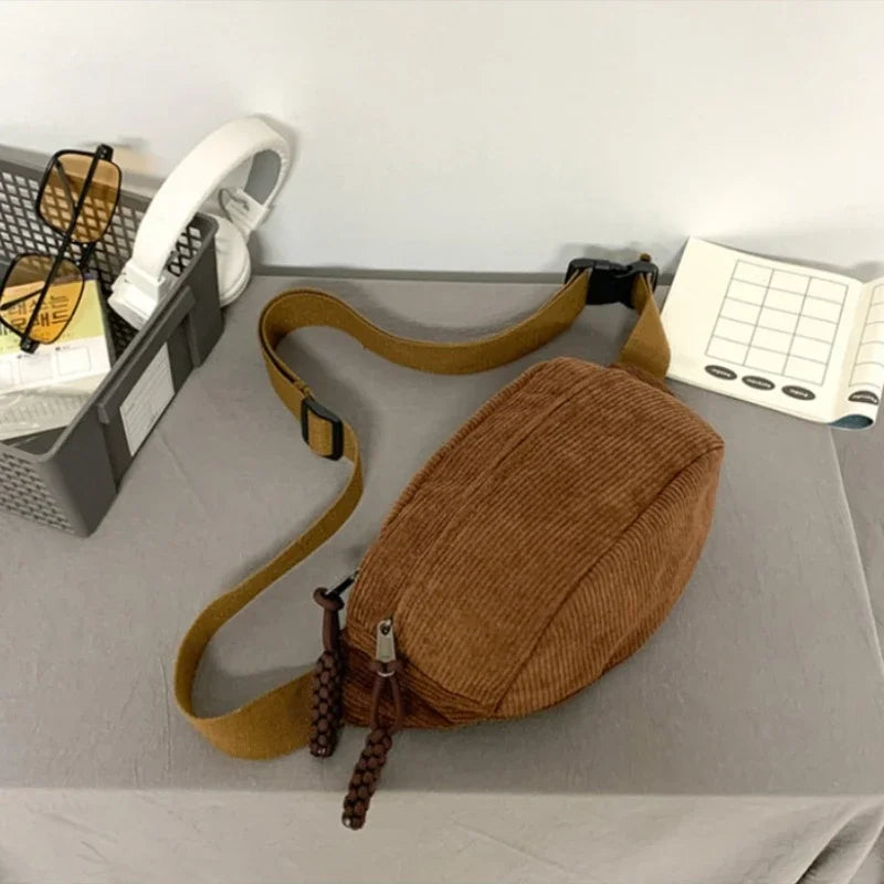 A brown corduroy waist pack for women is displayed on a gray surface next to some accessories and a notebook.