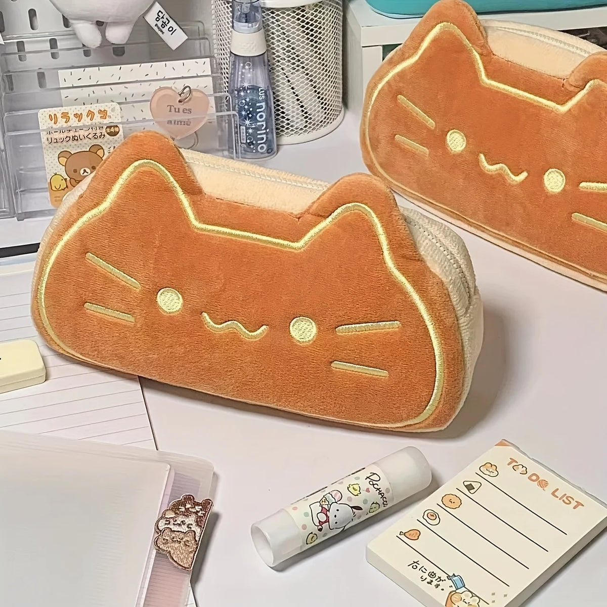 An orange caramel cat plush pencil pouch resting on a white surface next to other stationery items.