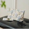A light blue floral canvas makeup bag with gold accents resting on a black leather surface.