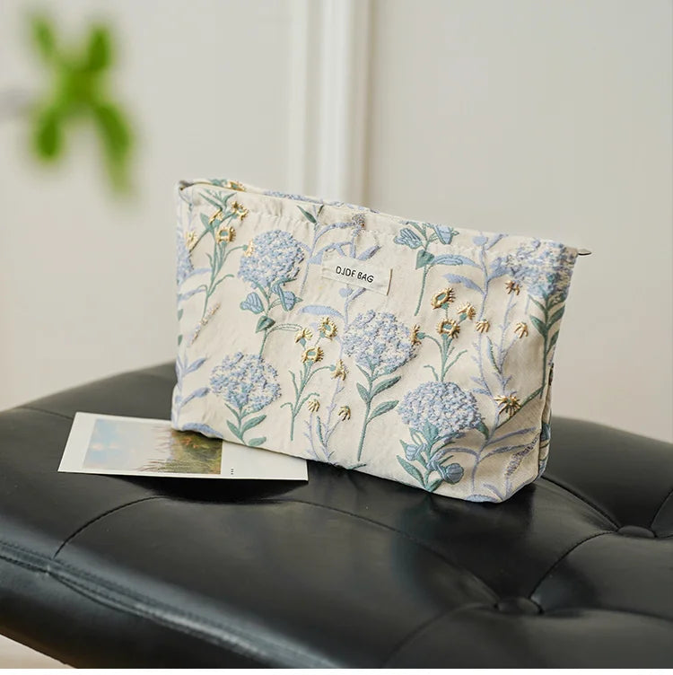 A light blue floral canvas makeup bag with gold accents resting on a black leather surface.