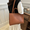 A person is holding a tan leather tote bag against a white paneled wall.