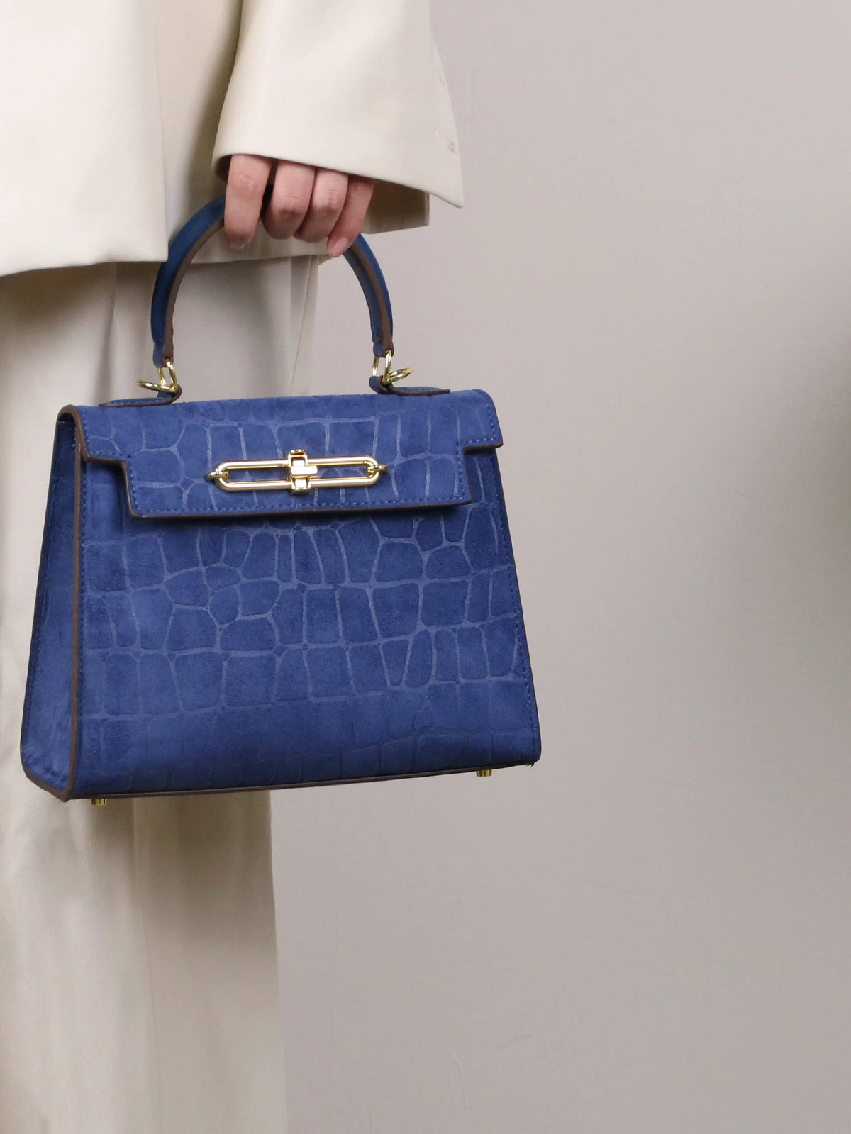 A navy blue crocodile pattern tote bag with gold hardware being held by a person in a light colored jacket.